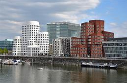 Das Foto zeigt die Gehry-Bauten im Düsseldorfer Medienhafen. Die drei markanten Gebäude mit geschwungenen Fassaden bestehen aus weißen, silbernen und roten Elementen und spiegeln sich im Wasser des Hafenbeckens. Im Vordergrund liegen Boote und ein größeres Schiff am Ufer.